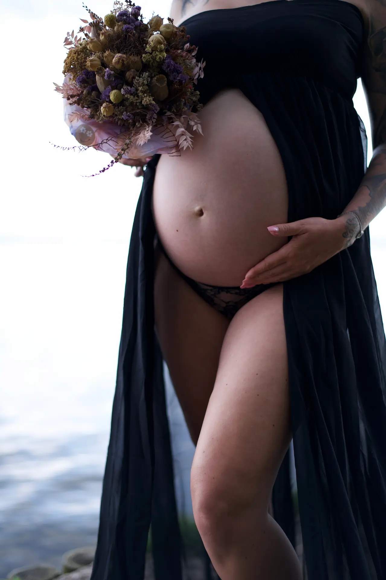 Babybauchfoto an einem Strand – Frau mit Blumenstrauß in der Hand.